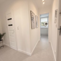 ITD. Usługi Budowlane - Jasny korytarz po remoncie z szarą podłogą, białymi ścianami, drzwiami z przeszkleniami i dekoracjami ściennymi w ramkach. W tle widoczna kuchnia.