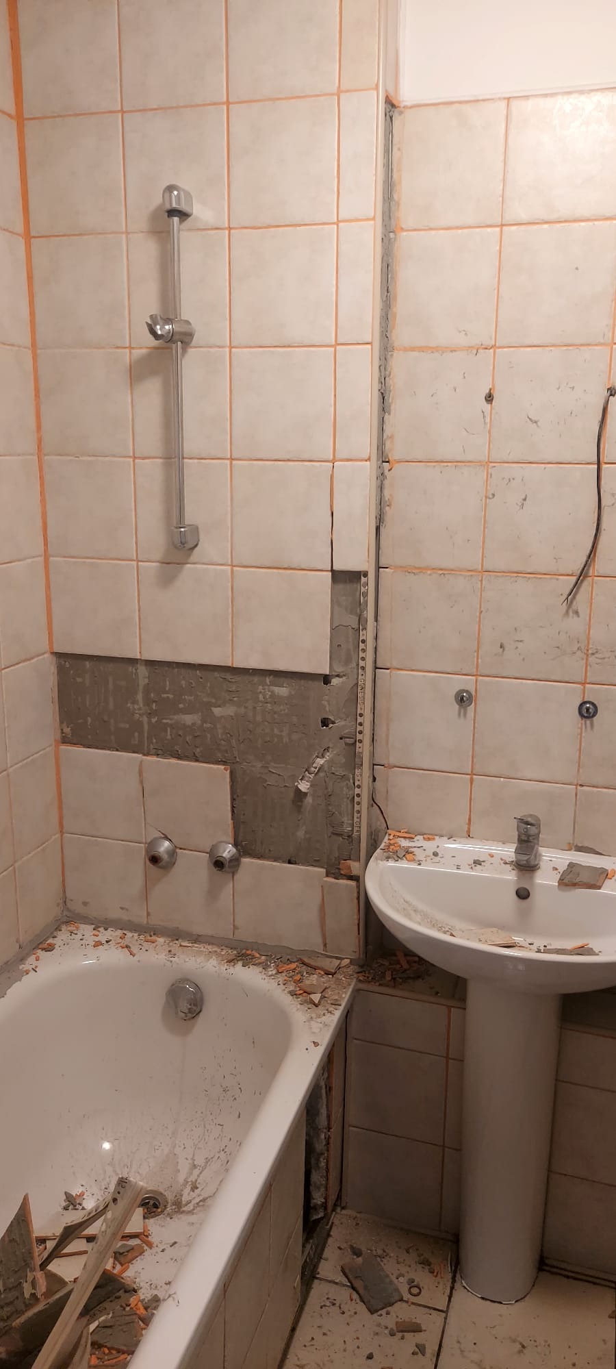 Demontaż płytek w łazience: fragment ściany bez glazury, widoczna instalacja, gruz na wannie i podłodze. Etap przygotowawczy do prac wykończeniowych.