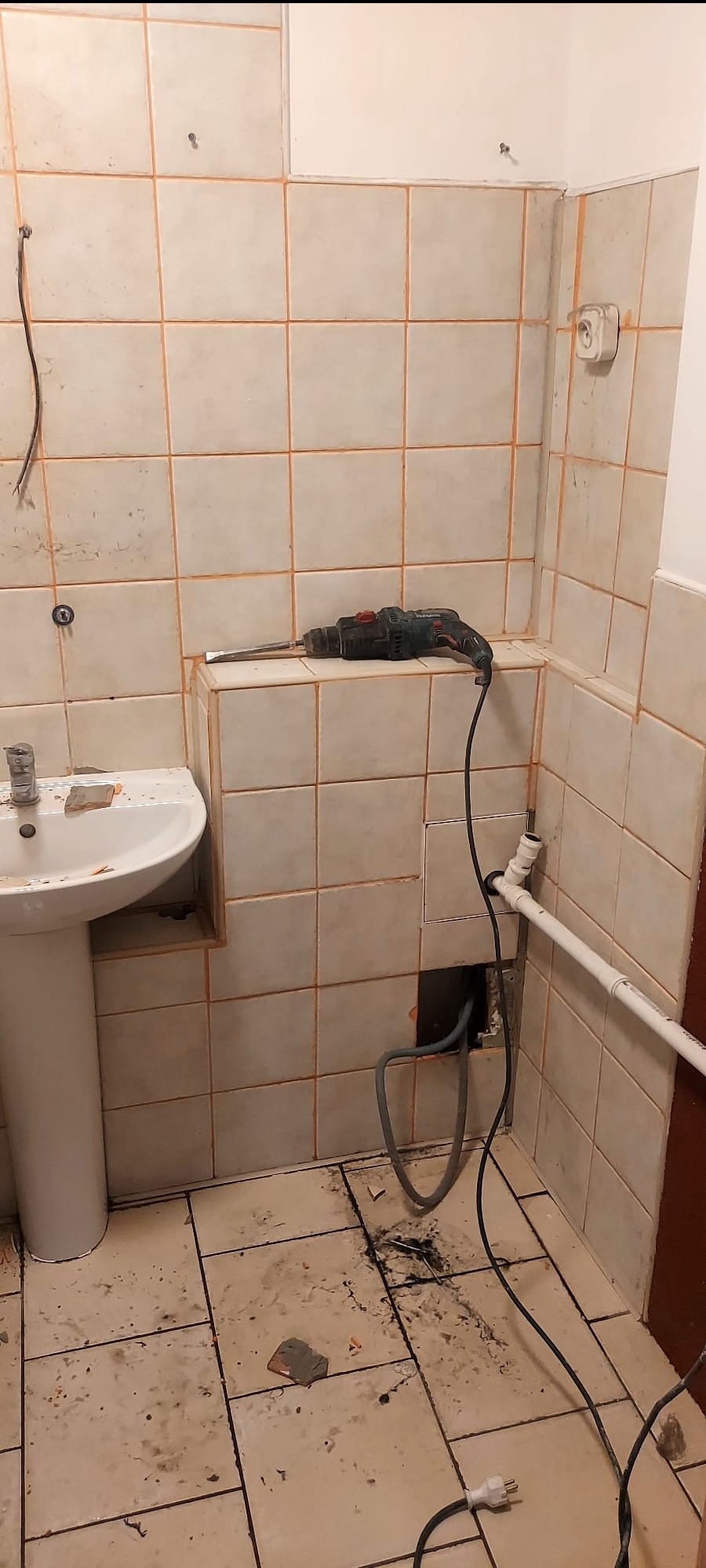 Demontaż płytek w łazience: widoczna umywalka, instalacja wodna, kable elektryczne i młot udarowy. Fragmenty płytek na podłodze.