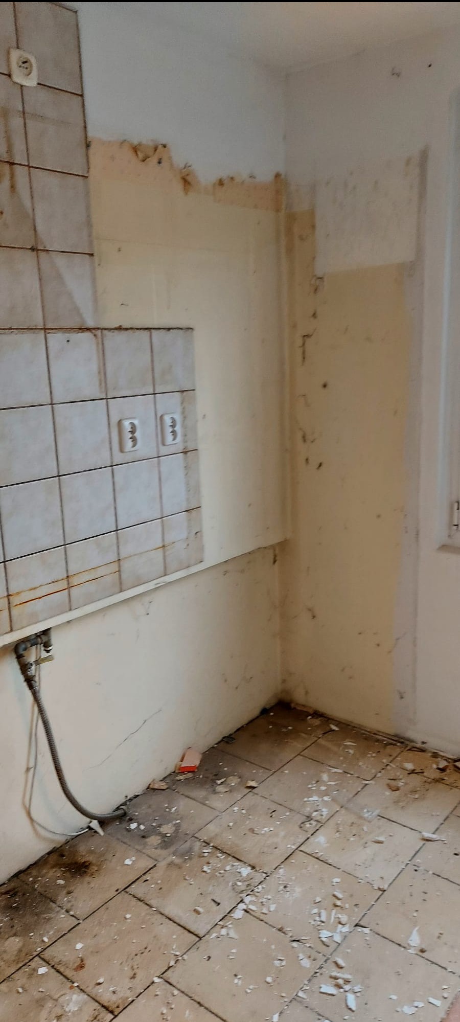 Kąt w kuchni w trakcie demontażu: stare płytki częściowo usunięte, odsłonięte gniazdka elektryczne, resztki kleju na ścianach, gruz na podłodze z płytek.