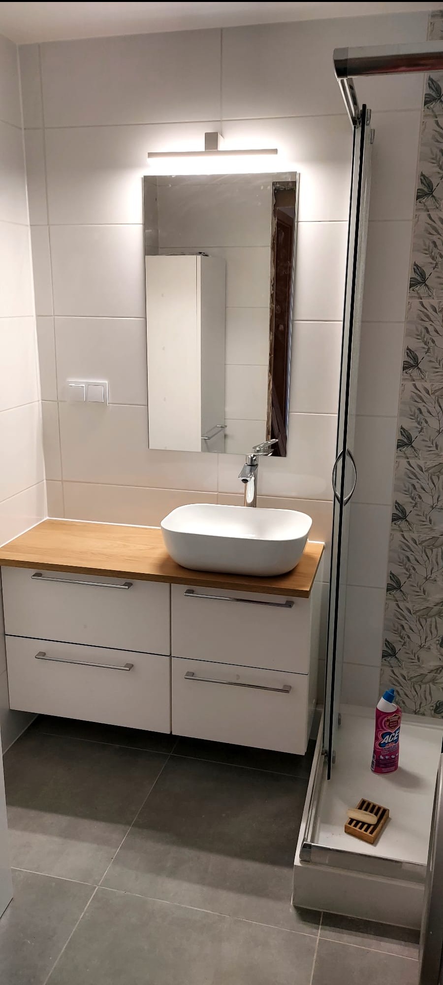 Nowoczesna łazienka: biała umywalka nablatowa na szafce z drewnianym blatem, lustro z oświetleniem, kabina prysznicowa z detalami i szare płytki podłogowe.