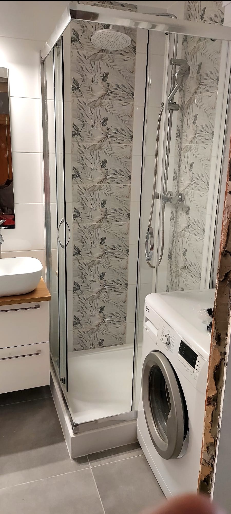 Wykończona łazienka z kabiną prysznicową z deszczownicą, umywalką na szafce i pralką. Na ścianach płytki z motywem liści. Widoczny fragment ściany w trakcie remontu.
