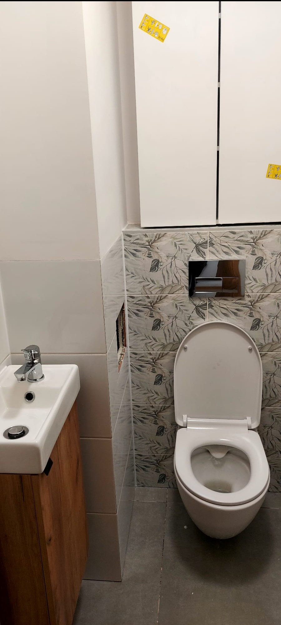 Wykończona łazienka: biała umywalka z drewnianą szafką, toaleta z zabudowaną spłuczką i płytki z motywem liści. Na ścianie naklejki informacyjne.