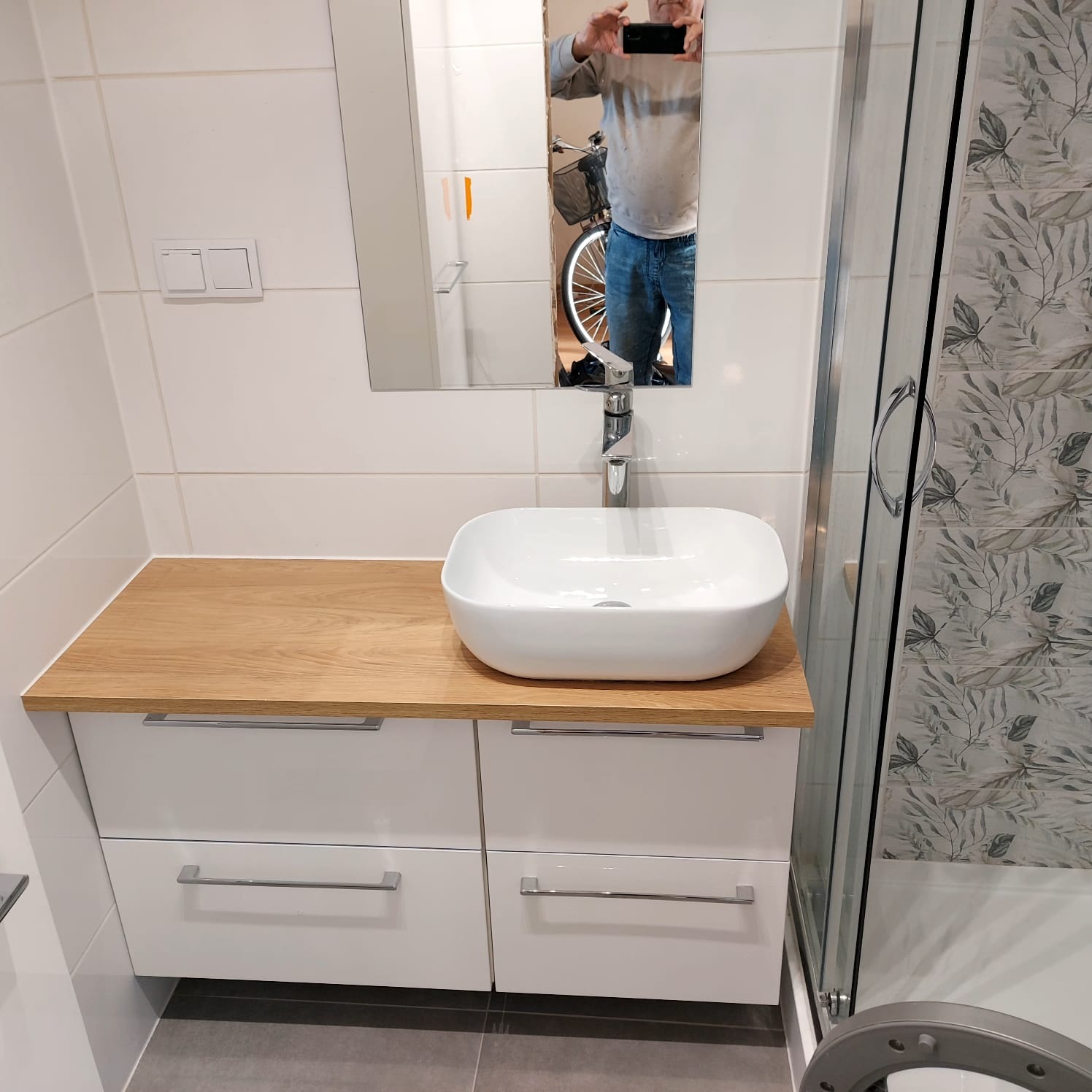 Nowoczesna łazienka: biała szafka z drewnianym blatem, umywalka nablatowa i kabina prysznicowa z roślinnym wzorem na płytkach. W lustrze odbicie osoby robiącej zdjęcie.