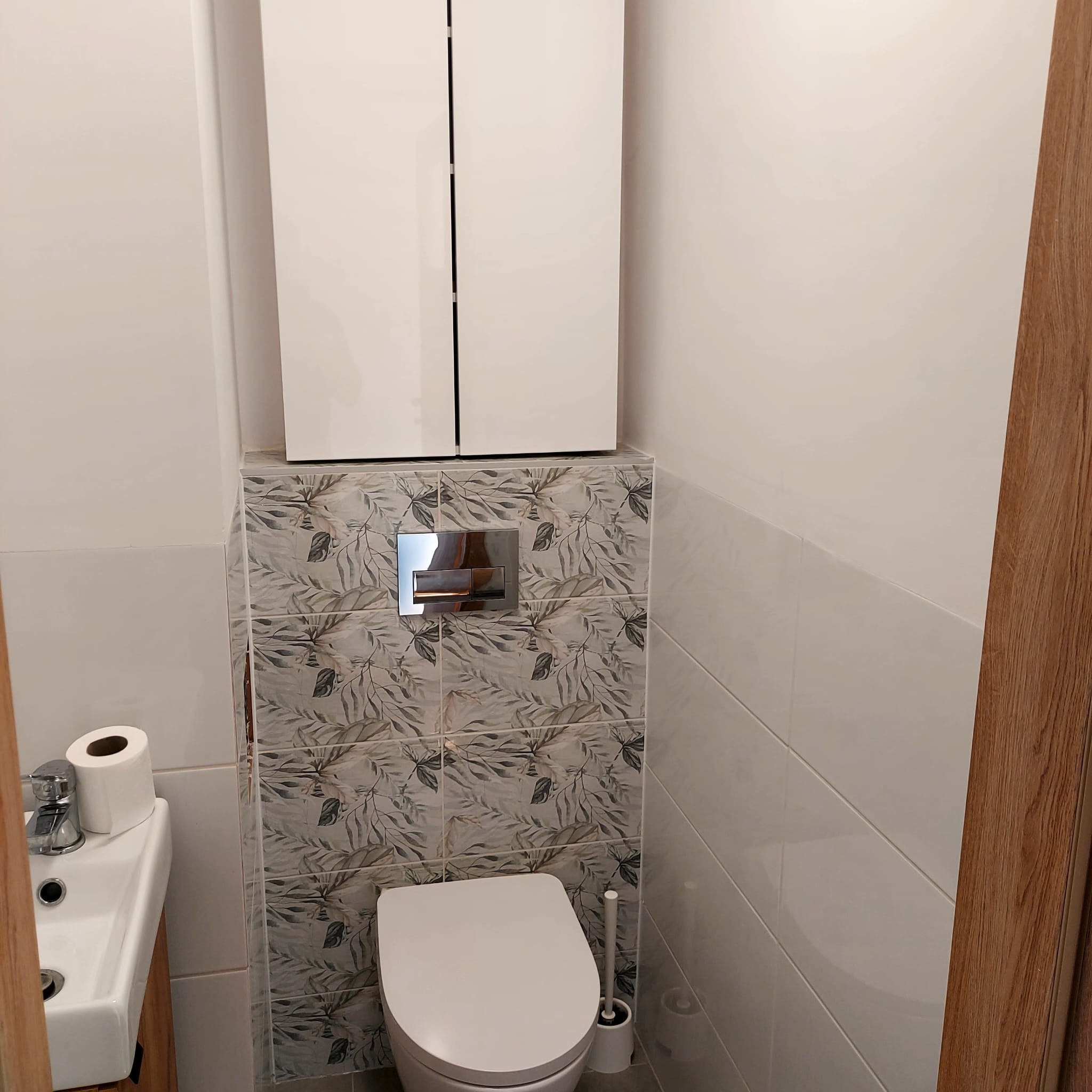 Wykończona łazienka: biała toaleta, szafka nad nią, umywalka z toaletą. Na ścianie płytki z motywem liści i chromowany przycisk spłuczki. Minimalistyczny design.