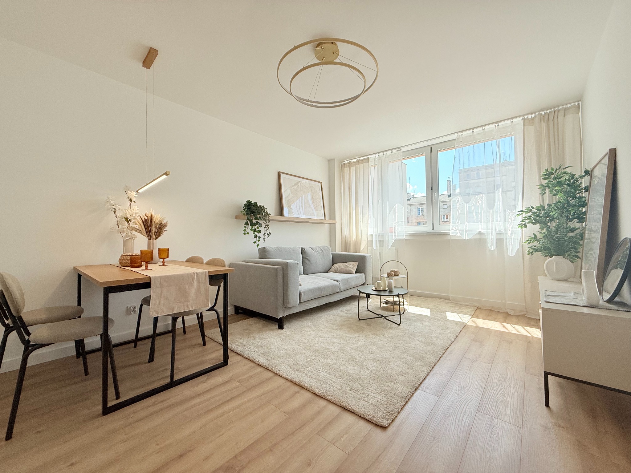 Jasne, przestronne wnętrze: stół z krzesłami, szara sofa na beżowym dywanie, okno z firankami. Całość w neutralnej kolorystyce z drewnianą podłogą.