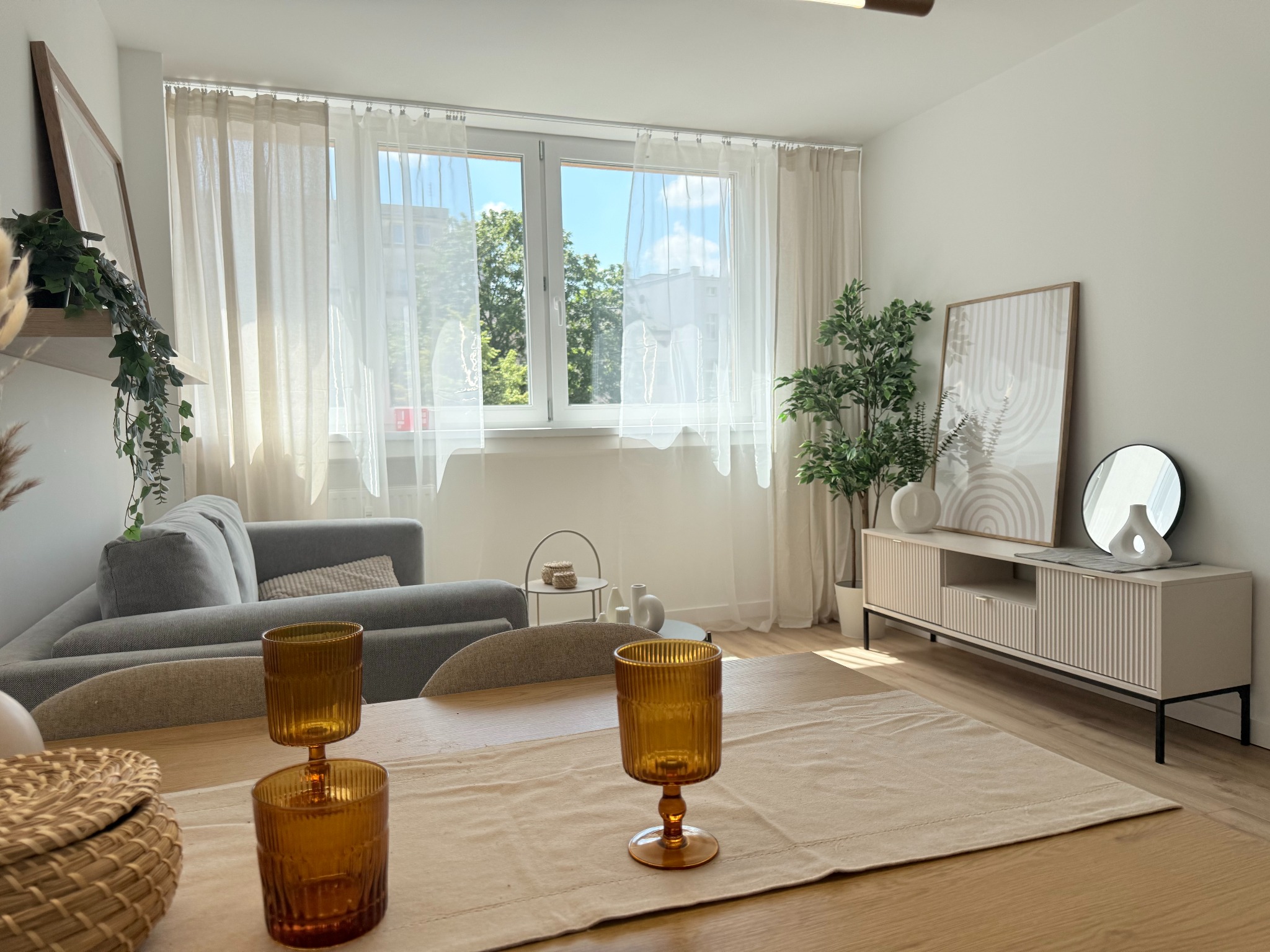 Jasne wnętrze salonu z drewnianym stołem i dekoracyjnymi kieliszkami. W tle szara sofa, okno z firanami i komoda z rośliną. Minimalistyczny, skandynawski styl.