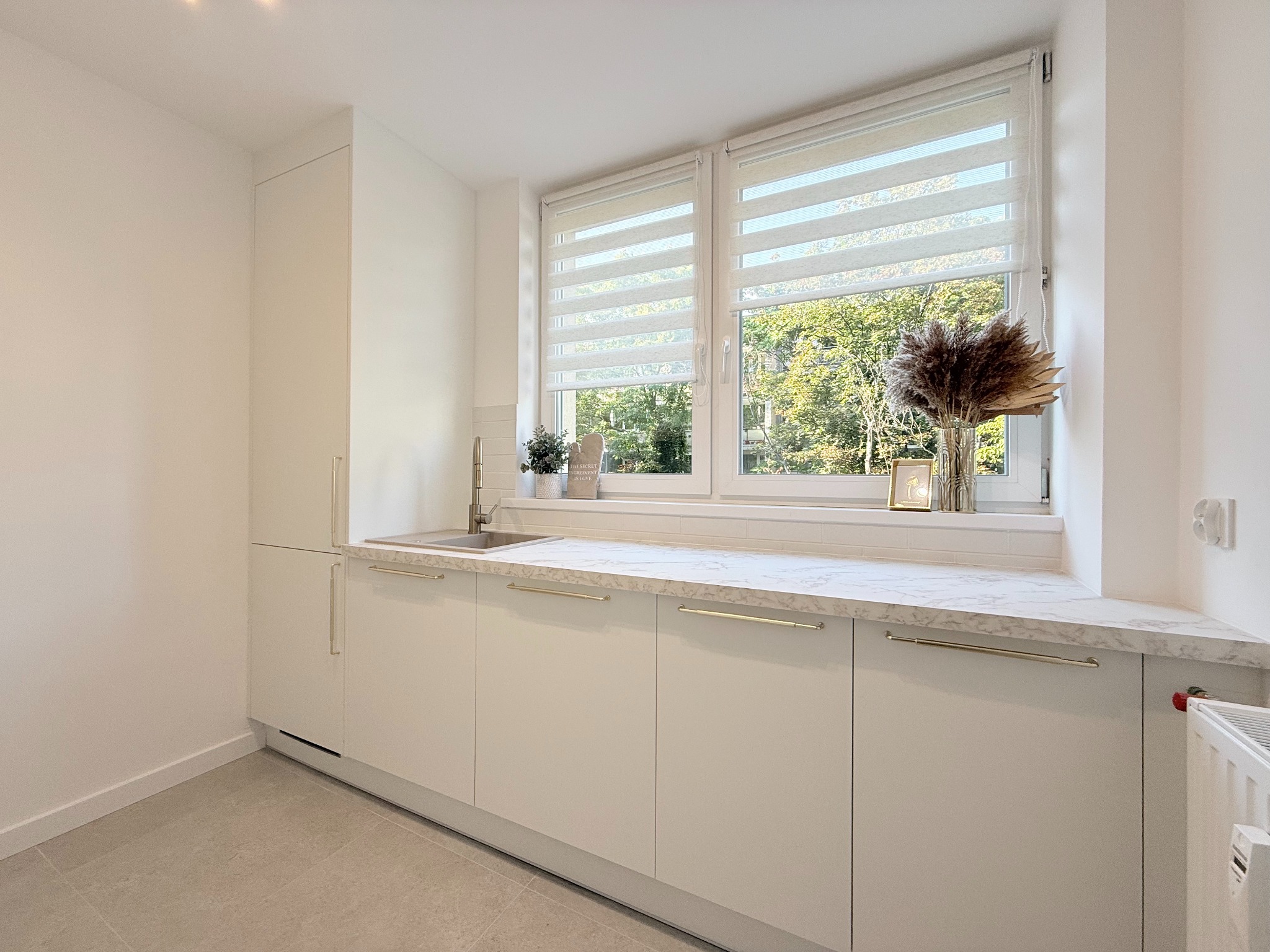 Jasna kuchnia z białymi szafkami, marmurowym blatem i oknem z roletami dzień-noc. Minimalistyczny design z dekoracjami na parapecie.