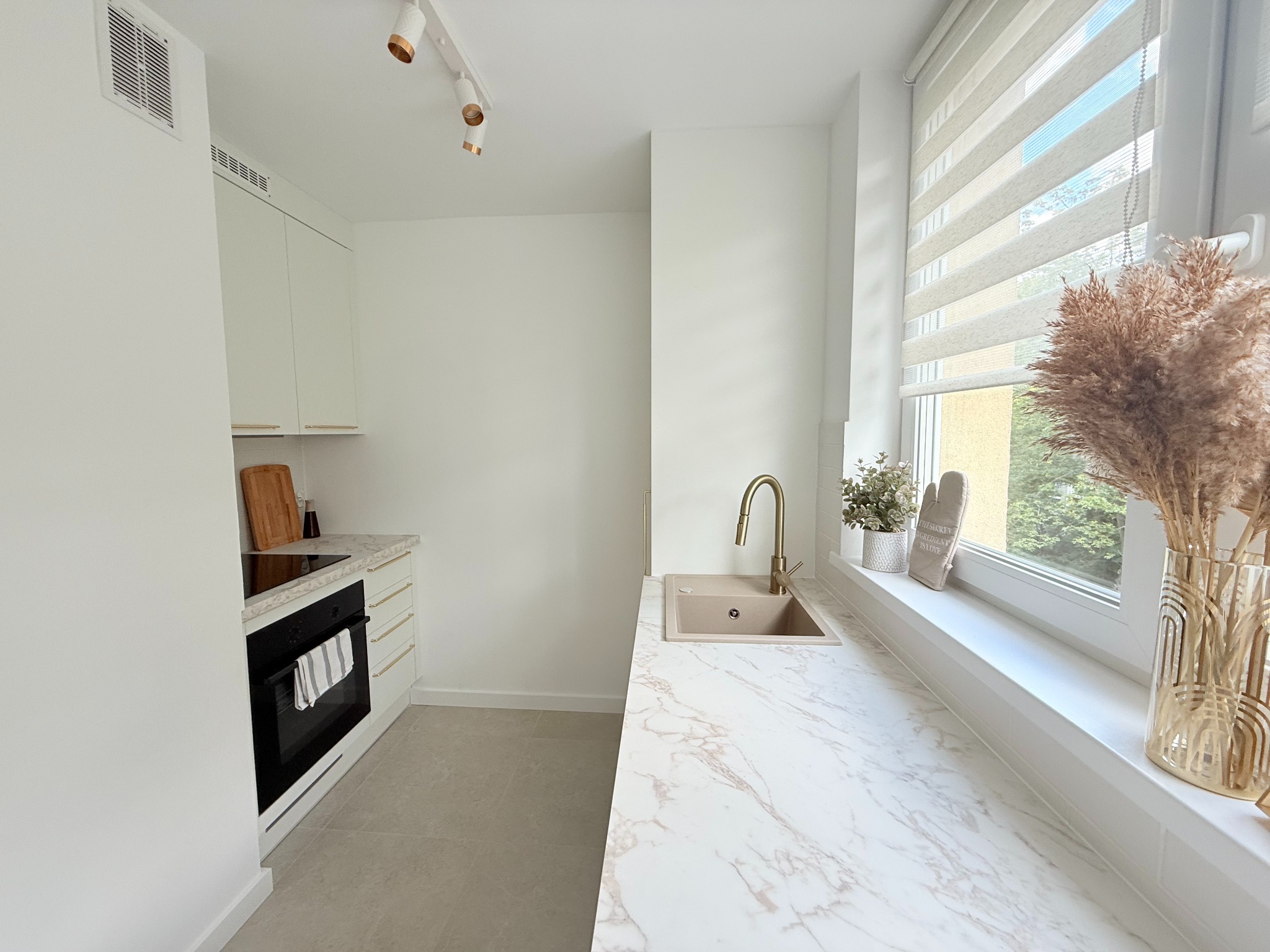 Jasna kuchnia z marmurowym blatem, złotą armaturą i dodatkami w stylu boho. Widoczne okno z roletami i dekoracje roślinne. Minimalistyczny design.