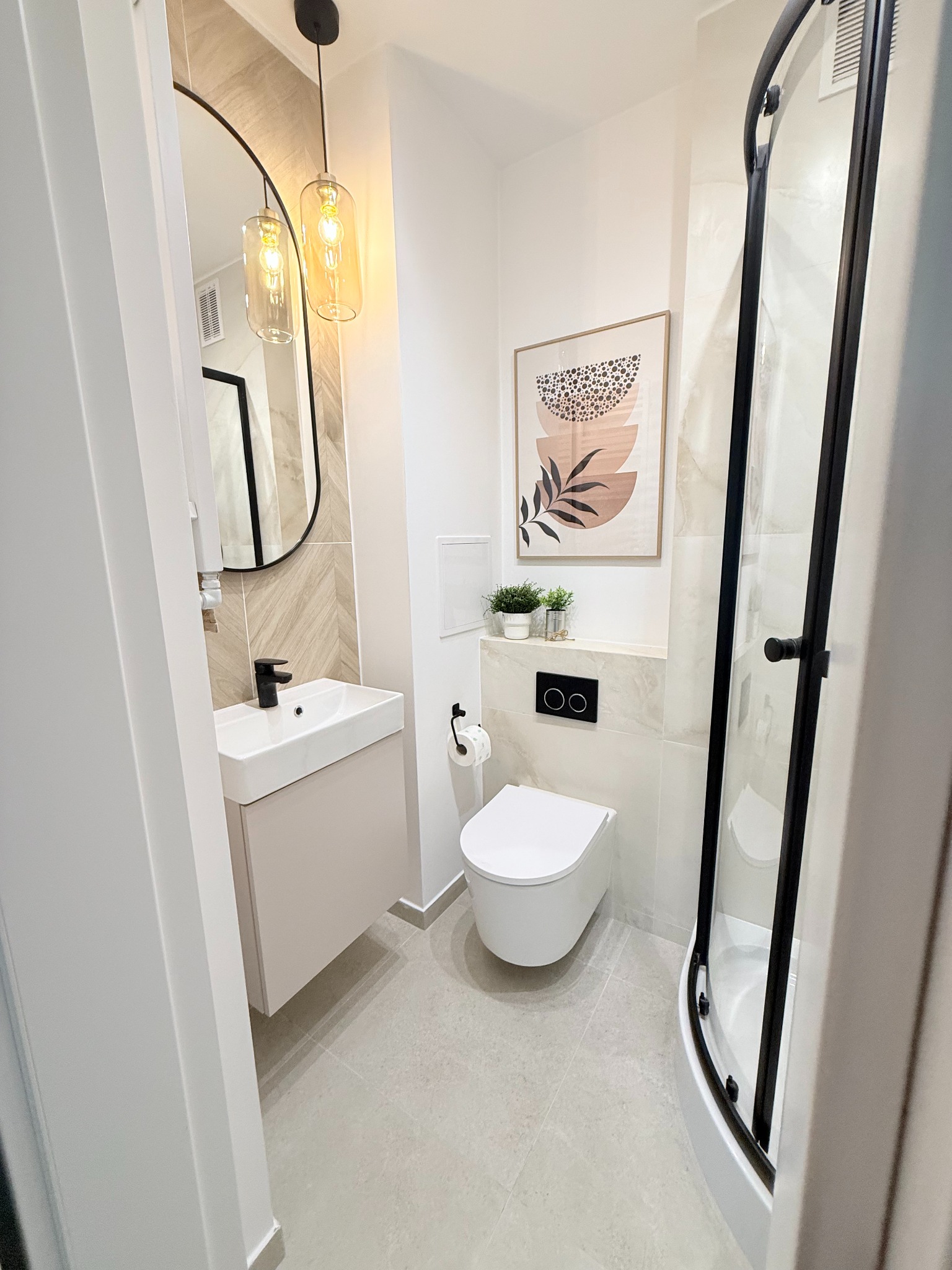 Nowoczesna łazienka w beżach: kabina prysznicowa z czarnymi ramami, umywalka z szafką, wisząca toaleta, lustro w czarnej ramie i grafika na ścianie.