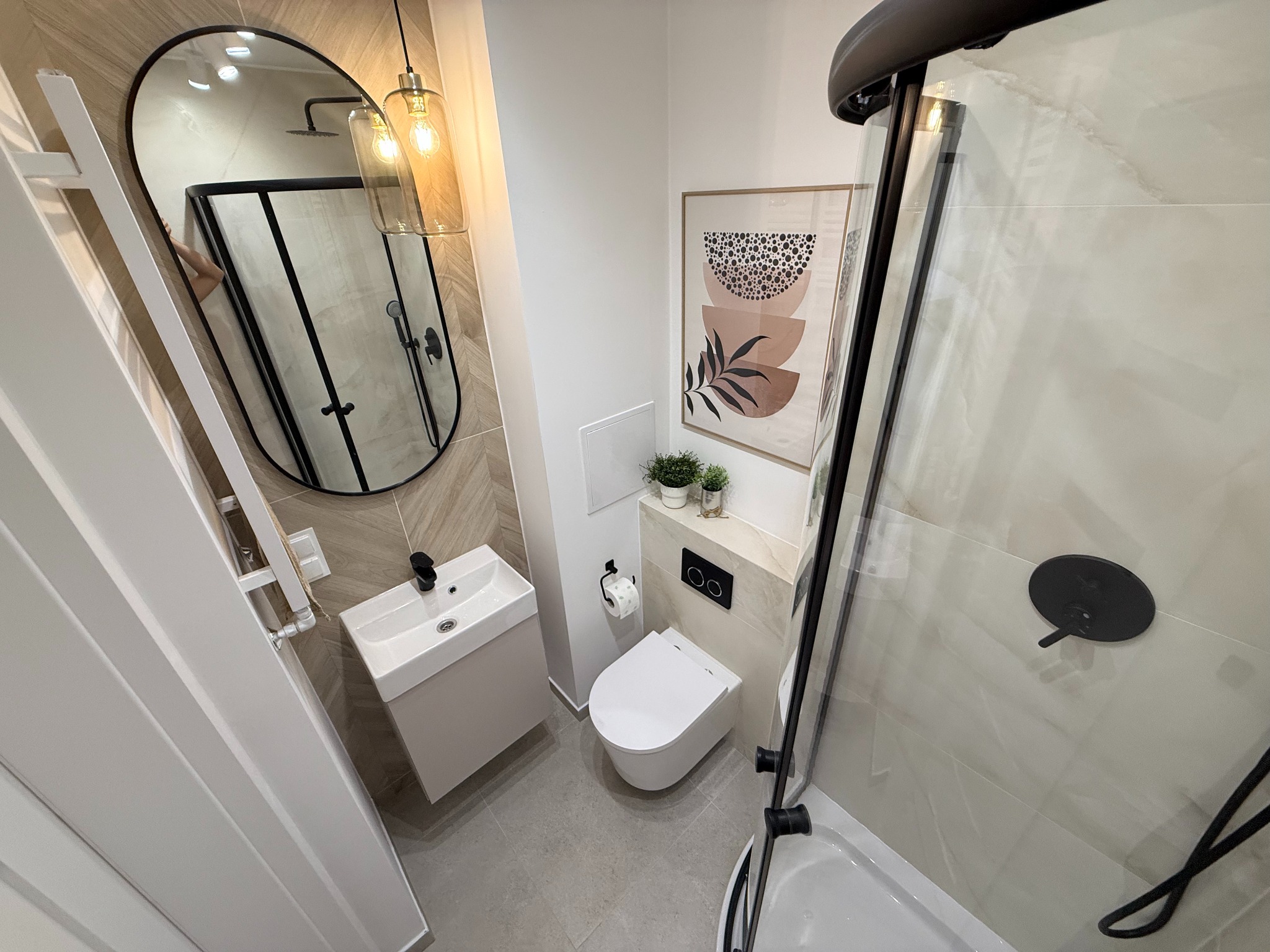Nowoczesna łazienka z owalnym lustrem, kabiną prysznicową z czarnymi detalami i umywalką. Na ścianie grafika w beżach i czerni, oświetlenie punktowe.