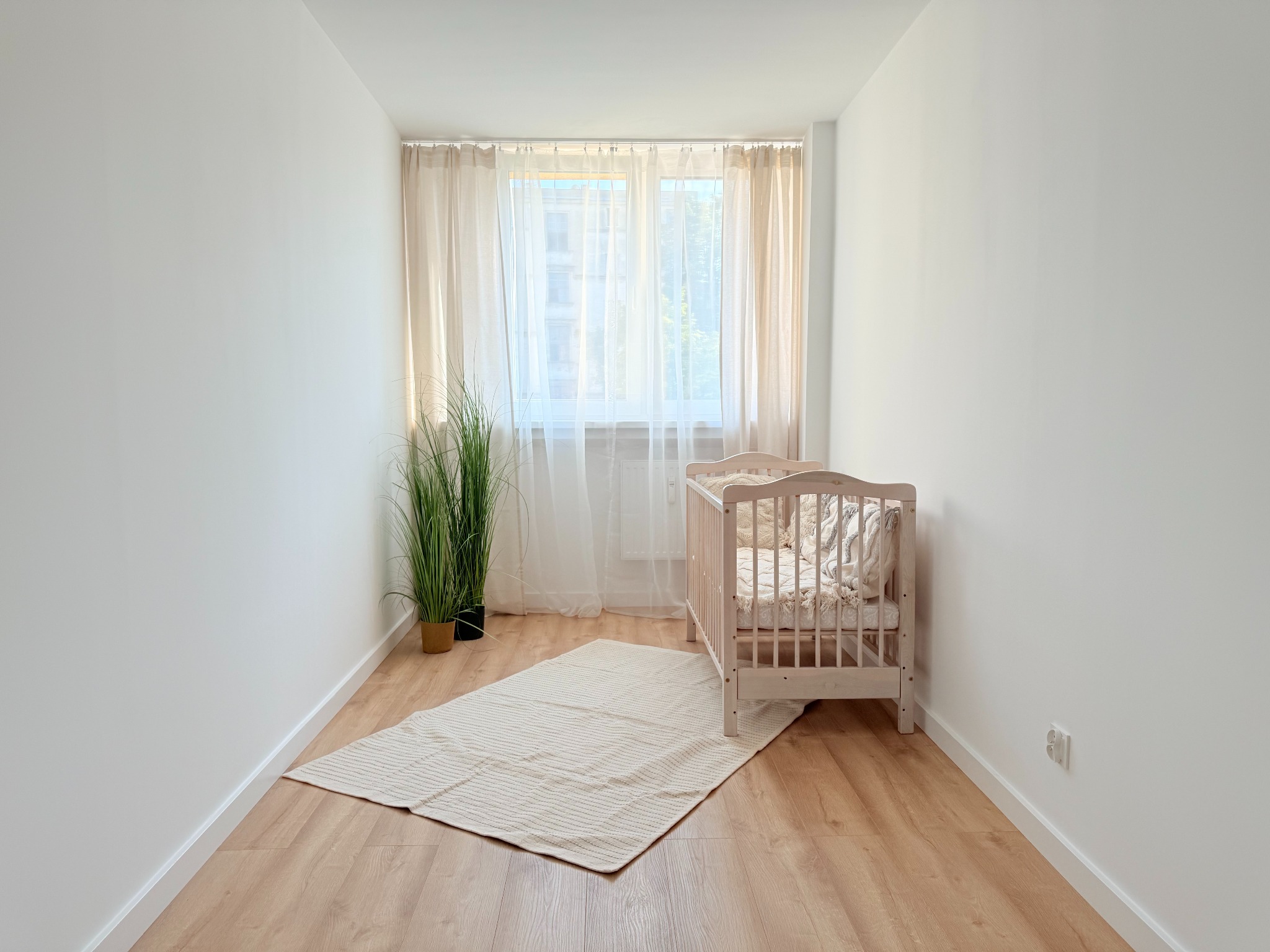 Jasne, minimalistyczne wnętrze pokoju dziecięcego w Oławie z drewnianym łóżeczkiem, dywanem i rośliną w doniczce przy oknie z beżowymi zasłonami.