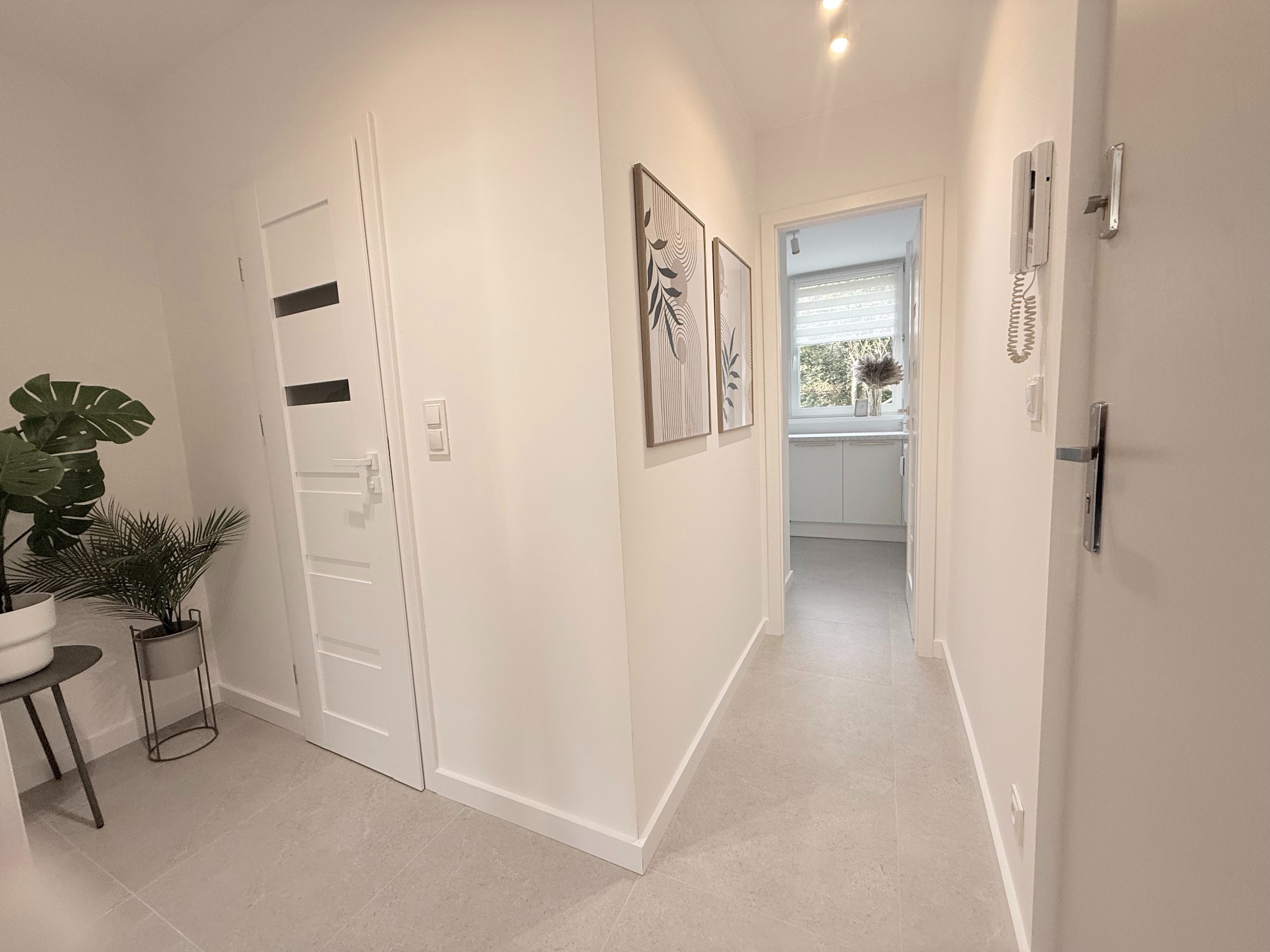 Jasny korytarz po remoncie z szarą podłogą, białymi ścianami, drzwiami z przeszkleniami i dekoracjami ściennymi w ramkach. W tle widoczna kuchnia.