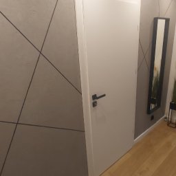 Łukasz Mikitów - Białe drzwi wewnętrzne z czarną klamką w nowoczesnym wnętrzu z szarą ścianą ozdobioną geometrycznym wzorem, drewniana podłoga, minimalistyczne oświetlenie sufitowe i lustro w czarnej ramie.