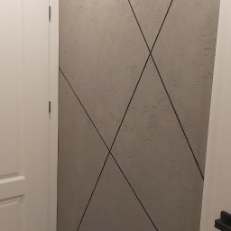 Łukasz Mikitów - Ściana pomalowana szarą farbą strukturalną z czarnymi, ukośnymi liniami dekoracyjnymi, widziana obok białych drzwi wewnętrznych i fragmentu podłogi z paneli.