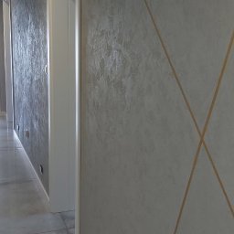 Łukasz Mikitów - Korytarz z szarą, teksturowaną ścianą ozdobioną złotymi liniami tworzącymi geometryczny wzór, białe futryny drzwiowe i szara podłoga z płytek.
