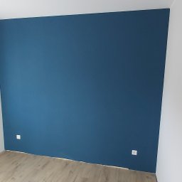 Łukasz Mikitów - Ściana pomalowana na ciemny niebieski kolor w Namysłowie, widoczne gniazdka elektryczne i fragment podłogi laminowanej.