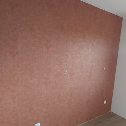 Łukasz Mikitów - Ściana w odcieniach brązu z widoczną strukturą, gniazdka elektryczne na dole, dwa uchwyty na ścianie, fragment podłogi.