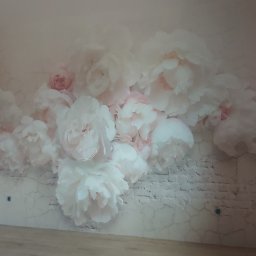 Łukasz Mikitów - Fototapeta z motywem białych piwonii na ścianie imitującej cegłę, widoczna podłoga i narzędzia malarskie w rogu.