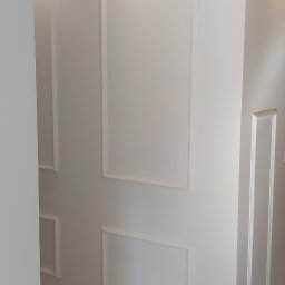 Łukasz Mikitów - Jasne wnętrze z dekoracyjnymi, prostokątnymi listwami ściennymi, oświetleniem punktowym i podłogą z paneli w odcieniach beżu.