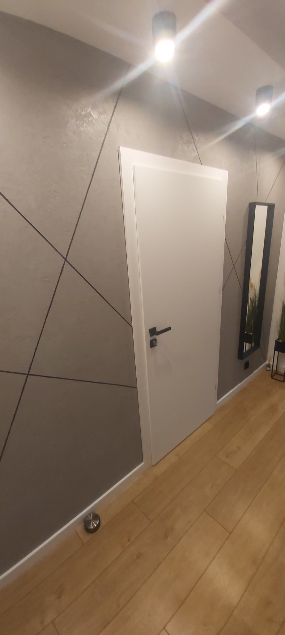 Białe drzwi wewnętrzne z czarną klamką w nowoczesnym wnętrzu z szarą ścianą ozdobioną geometrycznym wzorem, drewniana podłoga, minimalistyczne oświetlenie sufitowe i lustro w czarnej ramie.