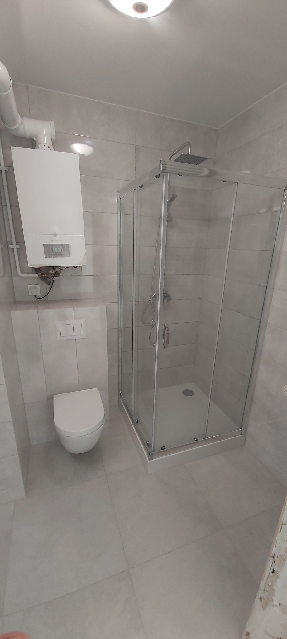 Nowoczesna łazienka z kabiną prysznicową, podwieszaną toaletą i piecem gazowym na ścianie, wykończona szarymi płytkami.