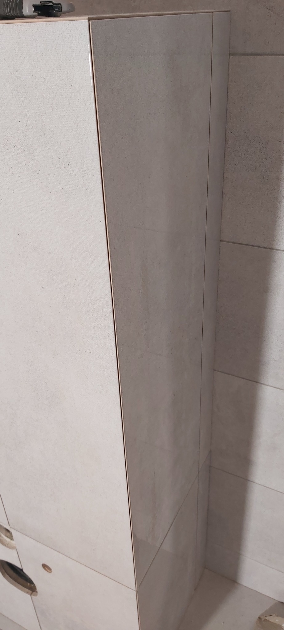 Narożnik obudowany płytkami ceramicznymi w łazience, widoczne łączenia płytek i fragment ściany obłożonej podobnymi płytkami, w górnym rogu widoczny nóż monterski.