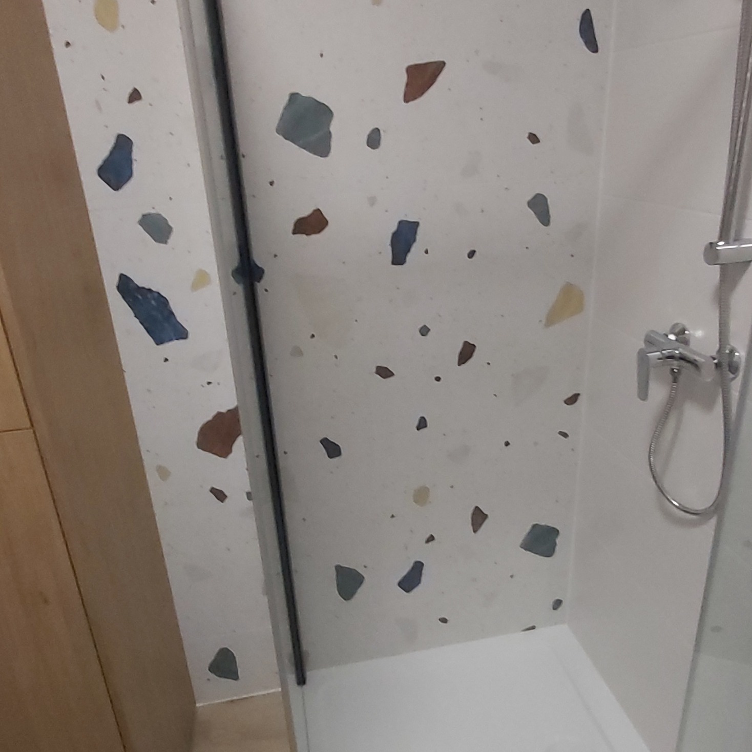 Biała ściana w kabinie prysznicowej z mozaiką w odcieniach niebieskiego, brązowego i żółtego, widoczna armatura prysznicowa i fragment drewnianej szafki.