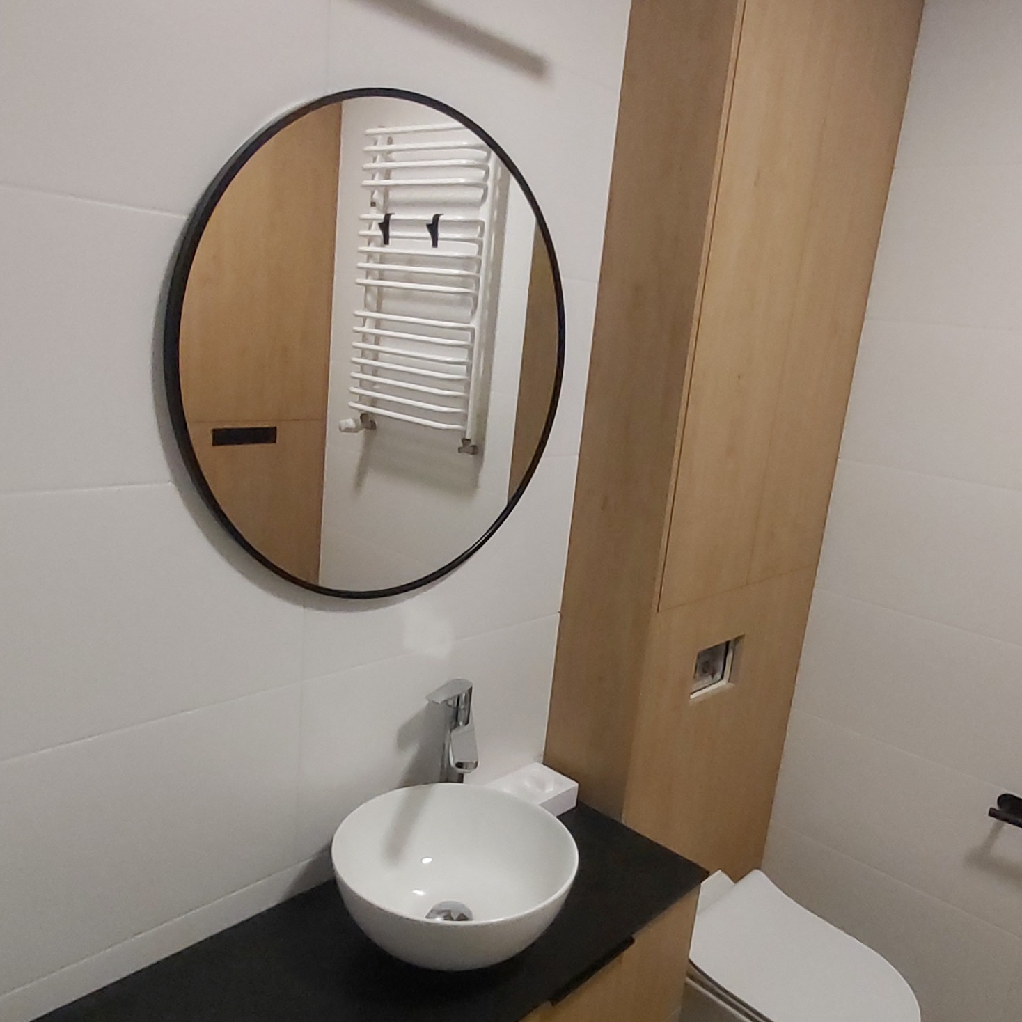 Nowoczesna łazienka z okrągłym lustrem, białą umywalką nablatową, drewnianą szafką i grzejnikiem drabinkowym widocznym w odbiciu lustra.
