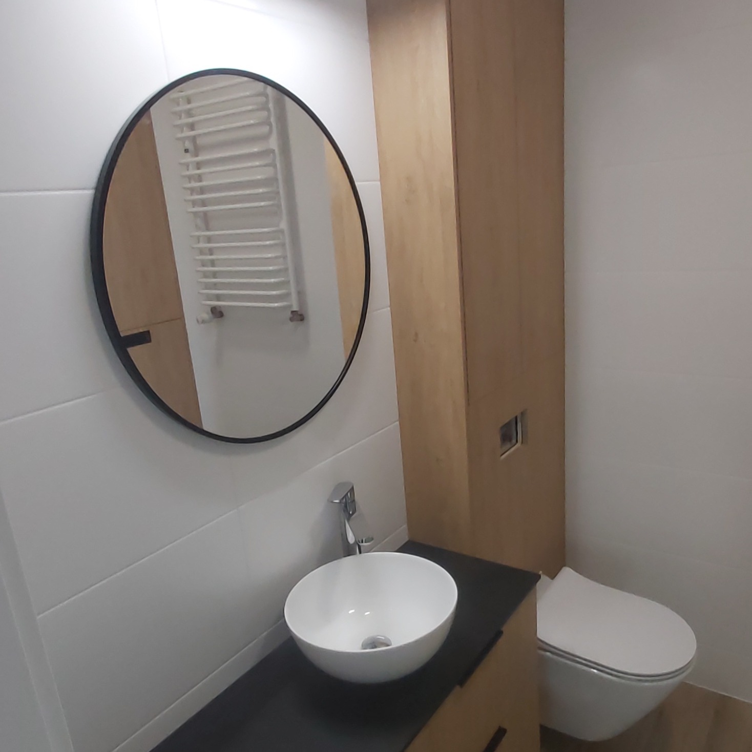 Nowoczesna łazienka z okrągłym lustrem z czarną ramą, białą umywalką nablatową, drewnianą szafką wiszącą i podwieszanym WC.