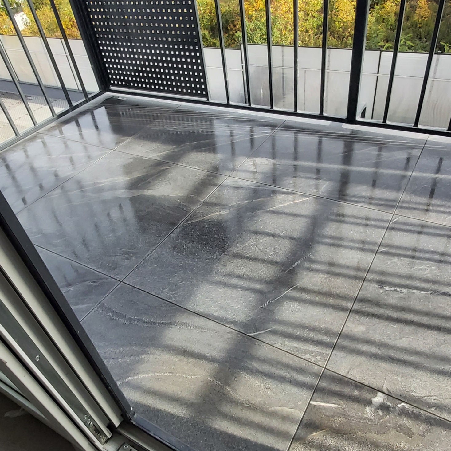 Błyszcząca, szara podłoga z płytek gresowych na balkonie z czarną, metalową balustradą i widokiem na drzewa.