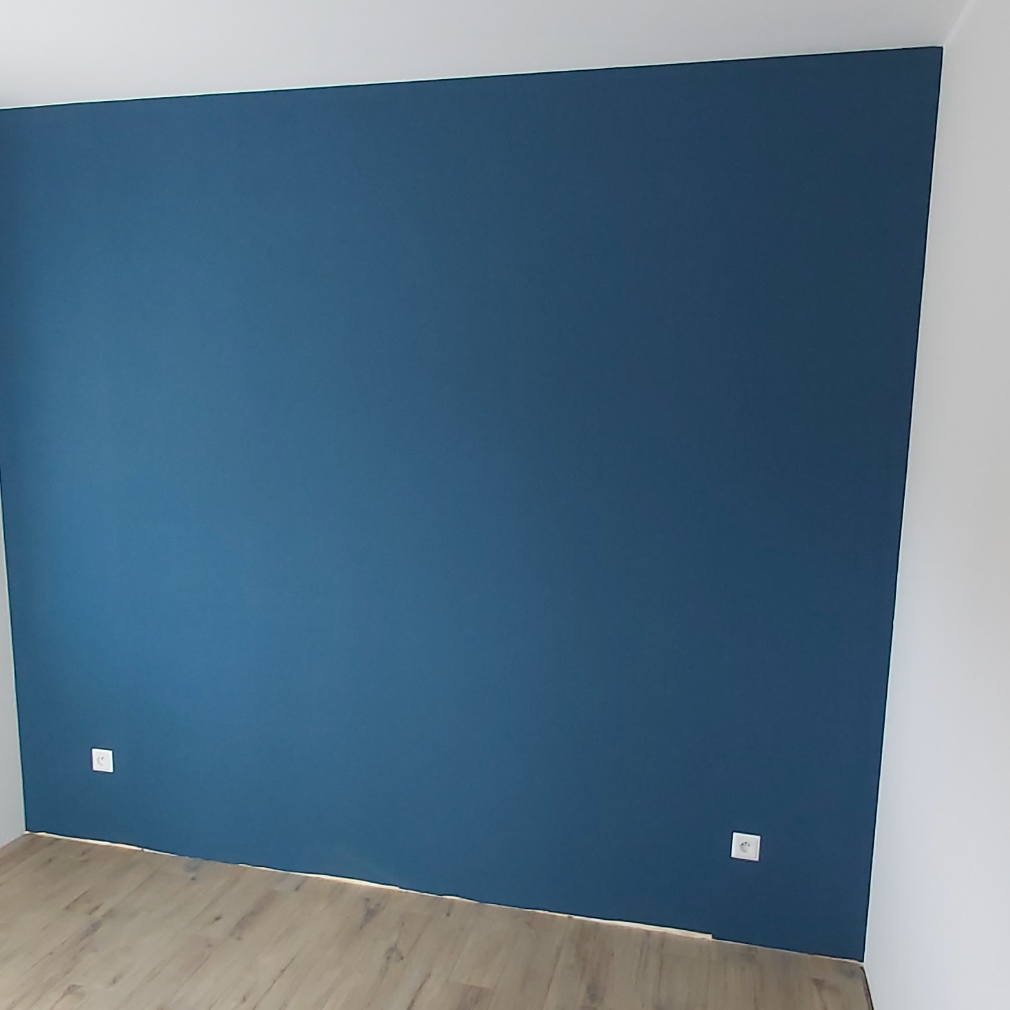 Ściana pomalowana na ciemny niebieski kolor w Namysłowie, widoczne gniazdka elektryczne i fragment podłogi laminowanej.