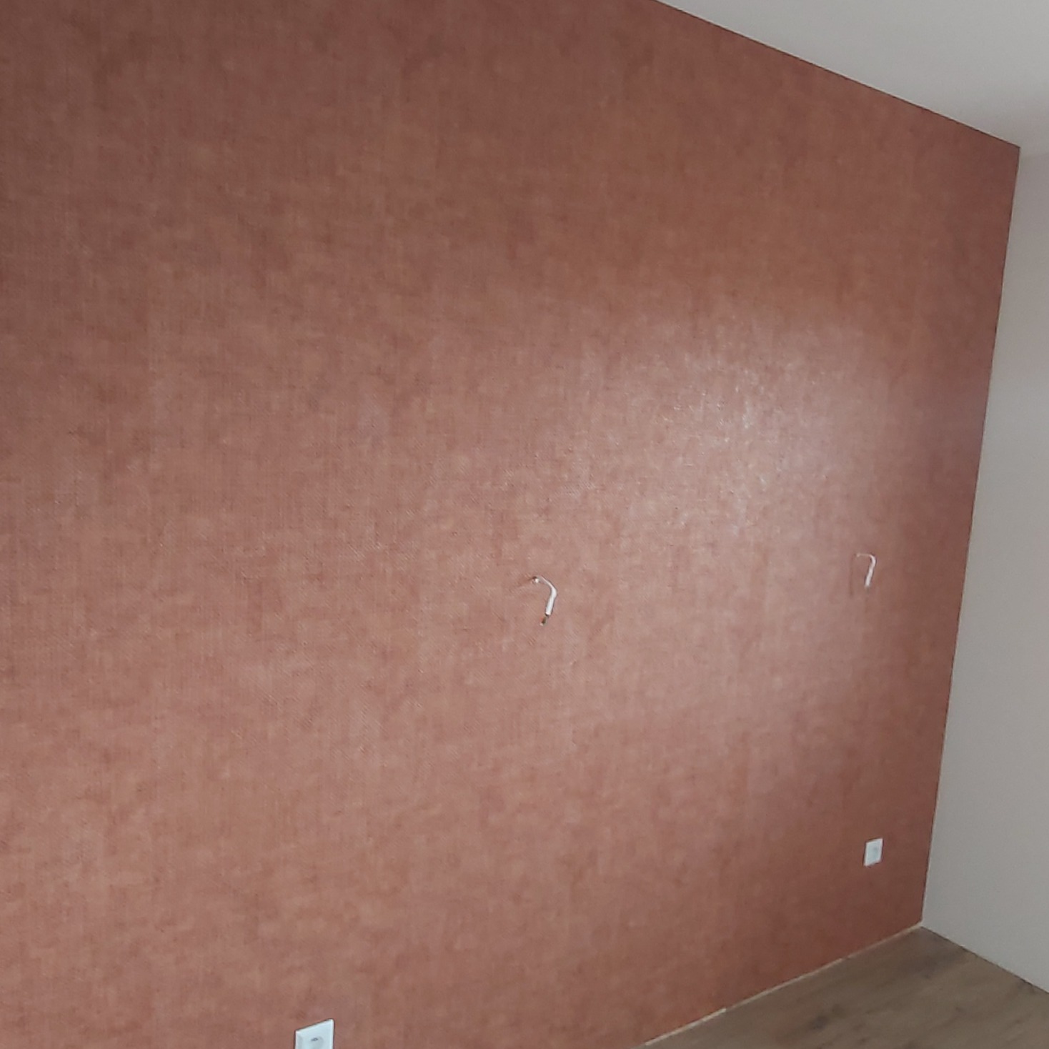 Ściana w odcieniach brązu z widoczną strukturą, gniazdka elektryczne na dole, dwa uchwyty na ścianie, fragment podłogi.