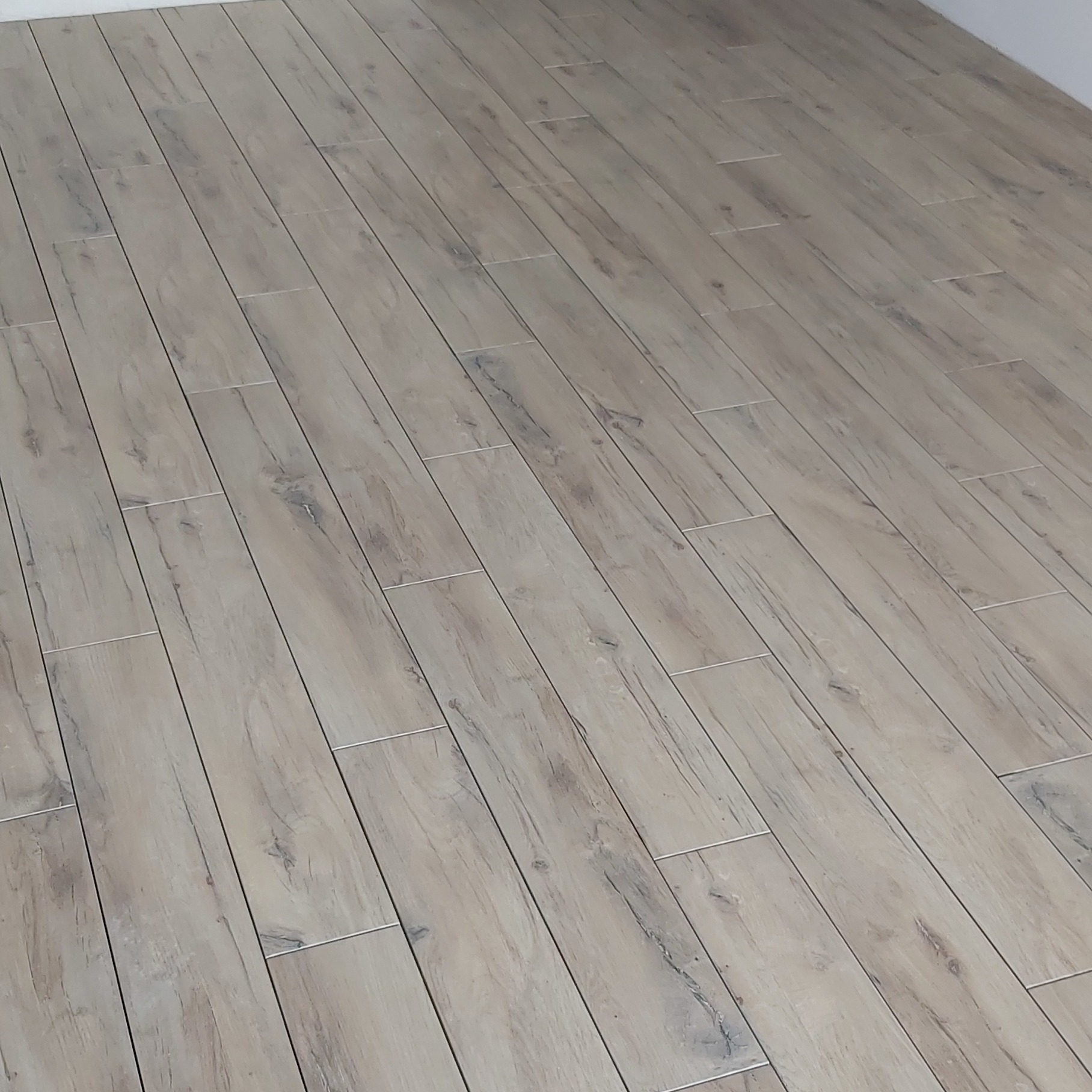 Ułożona pod kątem podłoga z płytek imitujących drewno w odcieniach beżu i szarości, z widocznymi fugami i fragmentem białej ściany w rogu.