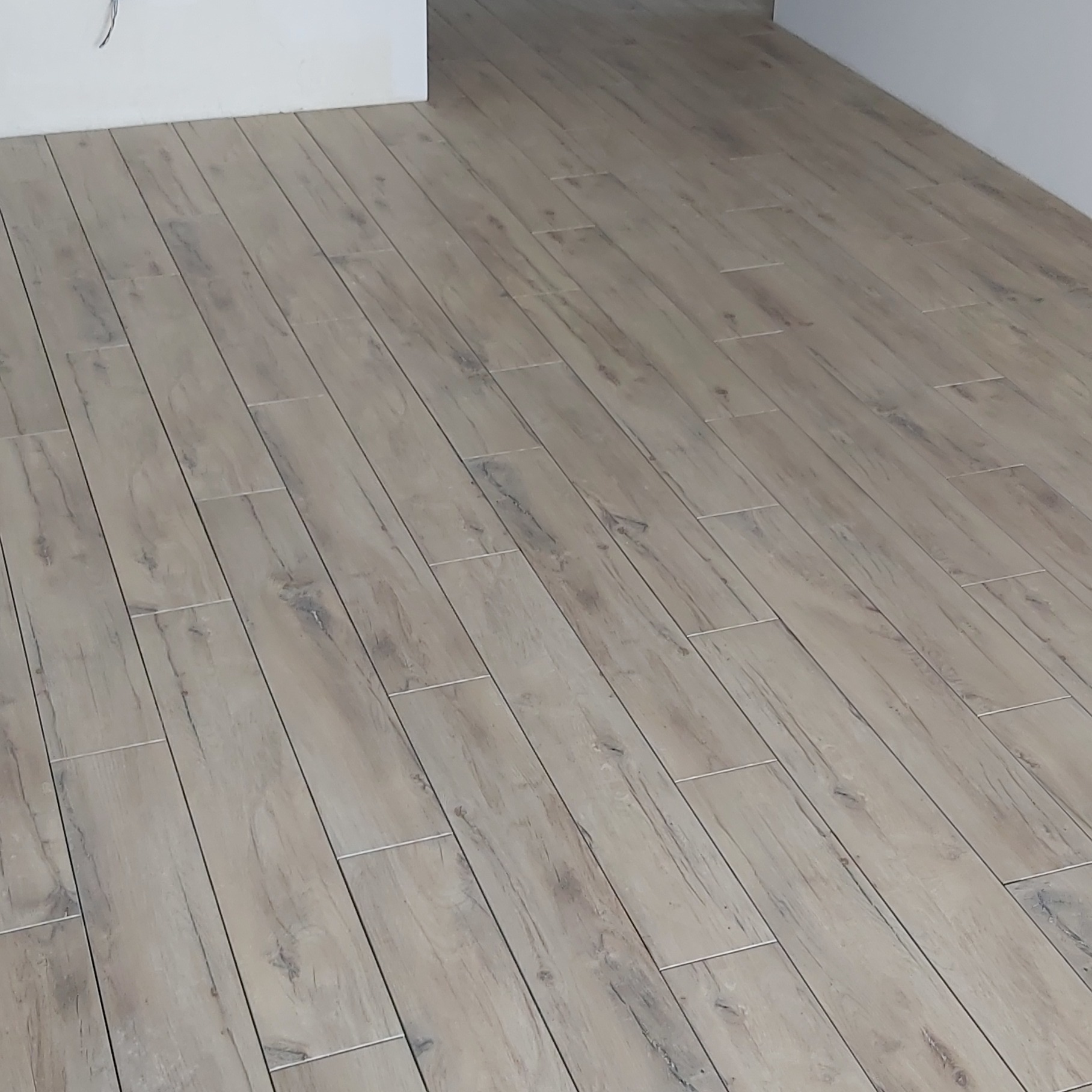Ułożona pod kątem podłoga z płytek imitujących drewno w odcieniach szarości, z widocznymi fugami i fragmentem białej ściany w tle.