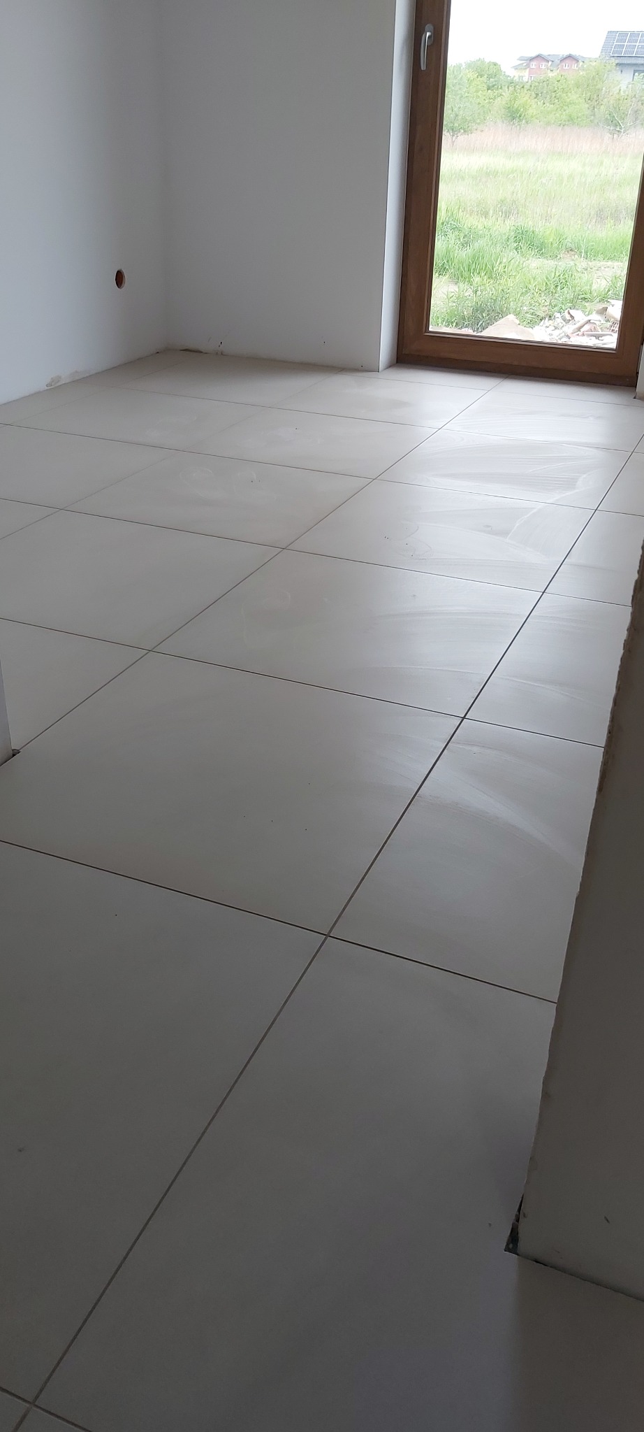 Ułożone, jasnoszare płytki podłogowe w nowym pomieszczeniu z oknem, widoczne ślady po fugowaniu.