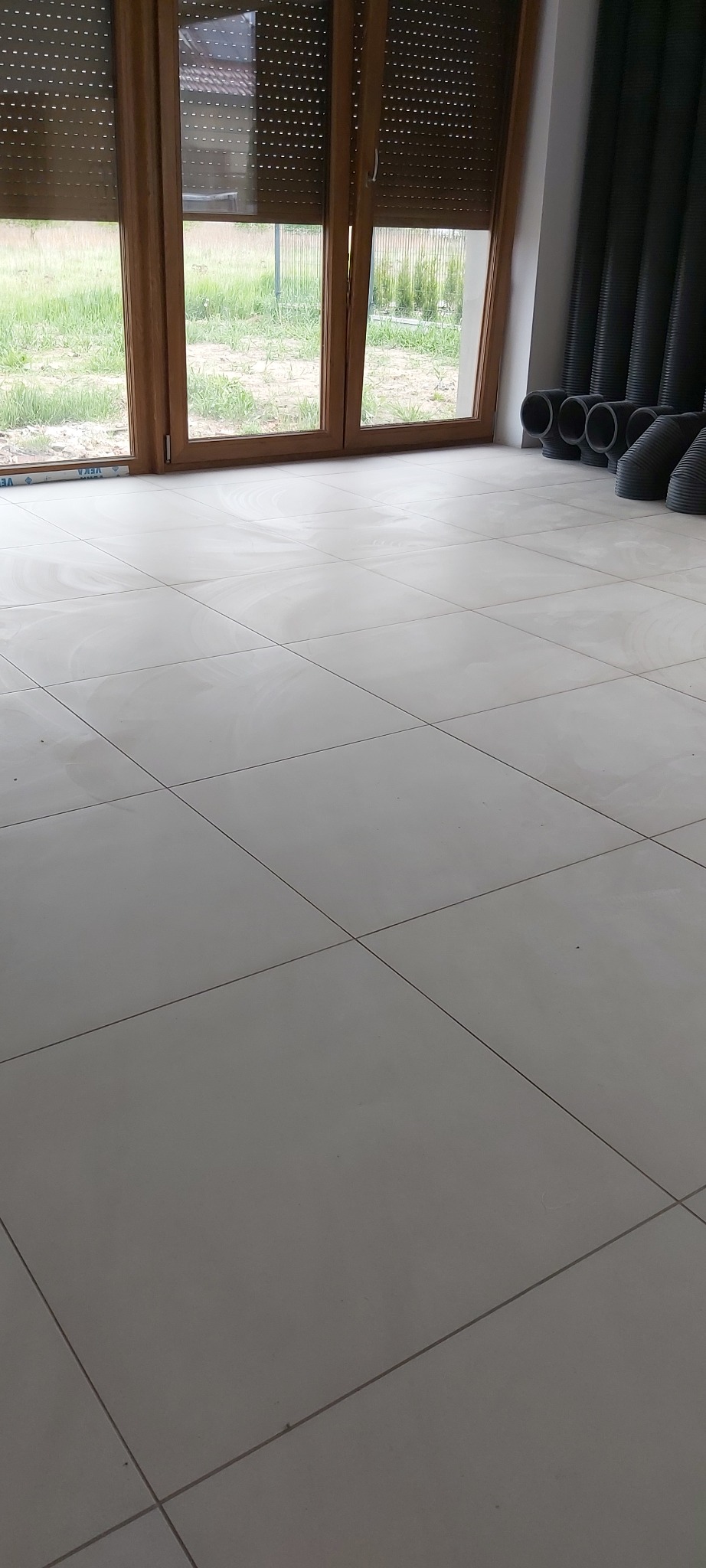 Jasne, kwadratowe płytki podłogowe z ciemnymi fugami, widoczne drewniane okno z roletami i czarne rury wentylacyjne w rogu pomieszczenia.