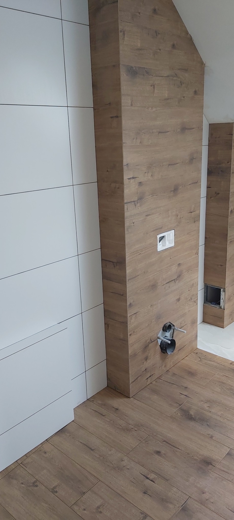 Nowoczesna łazienka w trakcie wykańczania, widoczne białe płytki na ścianie, obudowa z paneli drewnopodobnych z zamontowanym stelażem podtynkowym WC i instalacją hydrauliczną, oraz podłoga z paneli.