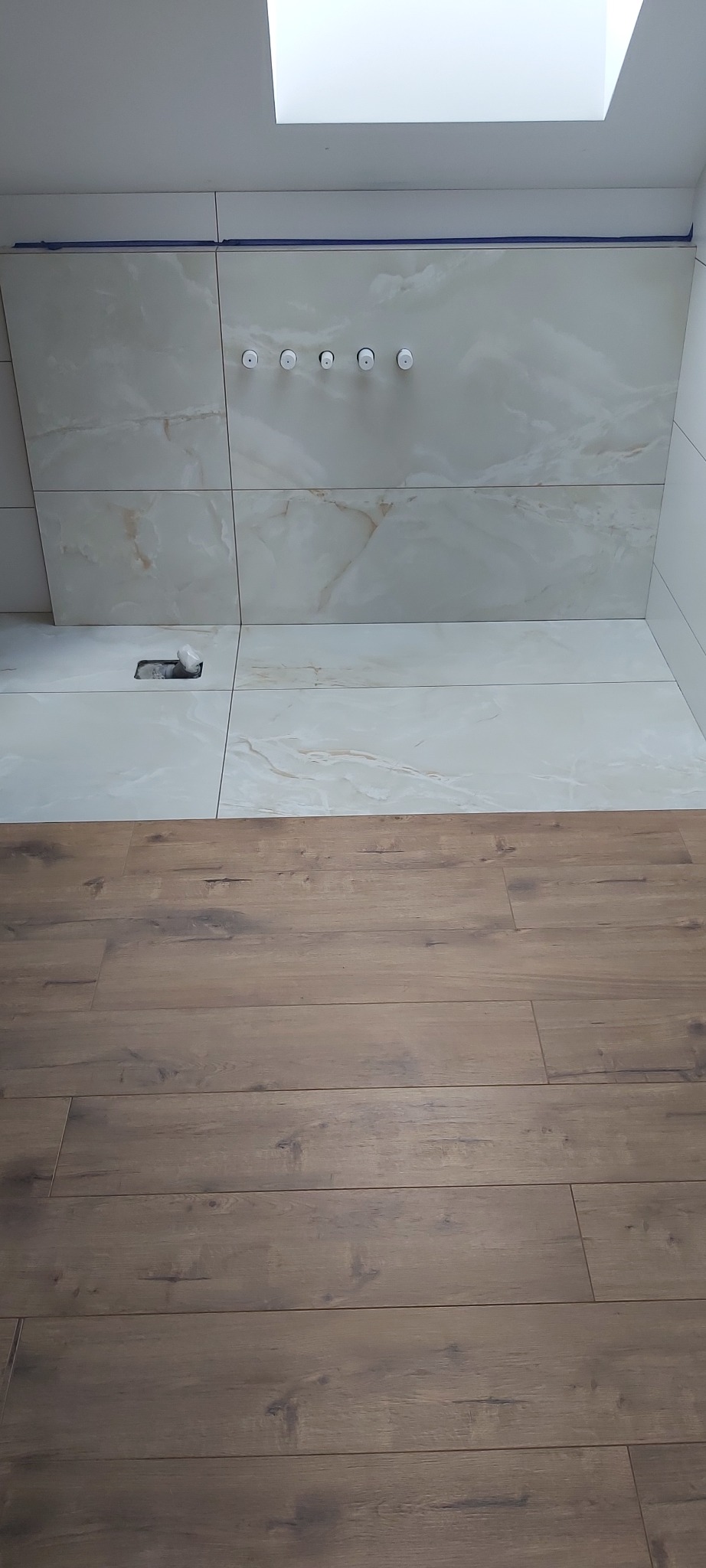 Ściana łazienki wyłożona jasnymi płytkami imitującymi marmur, z widocznymi otworami na armaturę i fragmentem podłogi z paneli drewnopodobnych, oświetlona światłem z okna dachowego.