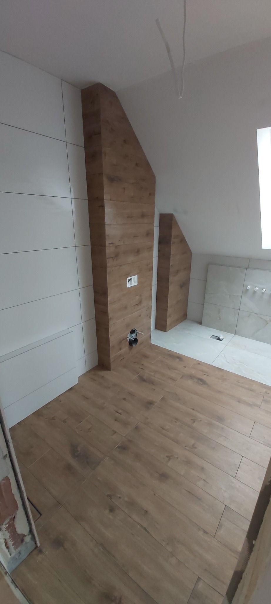 Wykończona łazienka na poddaszu z drewnopodobną podłogą i ścianami z białych płytek oraz zabudową z płytek drewnopodobnych wokół instalacji wodno-kanalizacyjnych.