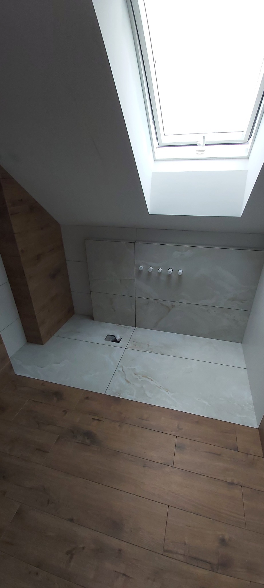 Ukończona kabina prysznicowa w skosie poddasza z oknem dachowym, wyłożona jasnymi płytkami imitującymi marmur, z widocznymi przyłączami do armatury i odpływem liniowym.