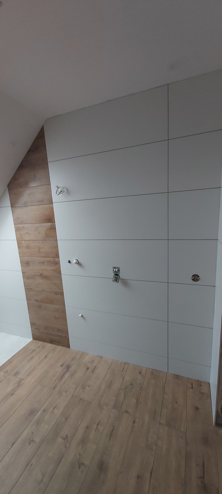 Ściana w łazience z poziomymi, białymi płytkami i dekoracyjnym pasem płytek drewnopodobnych, widoczne przygotowane punkty instalacji wodnej i elektrycznej, na podłodze panele drewnopodobne.