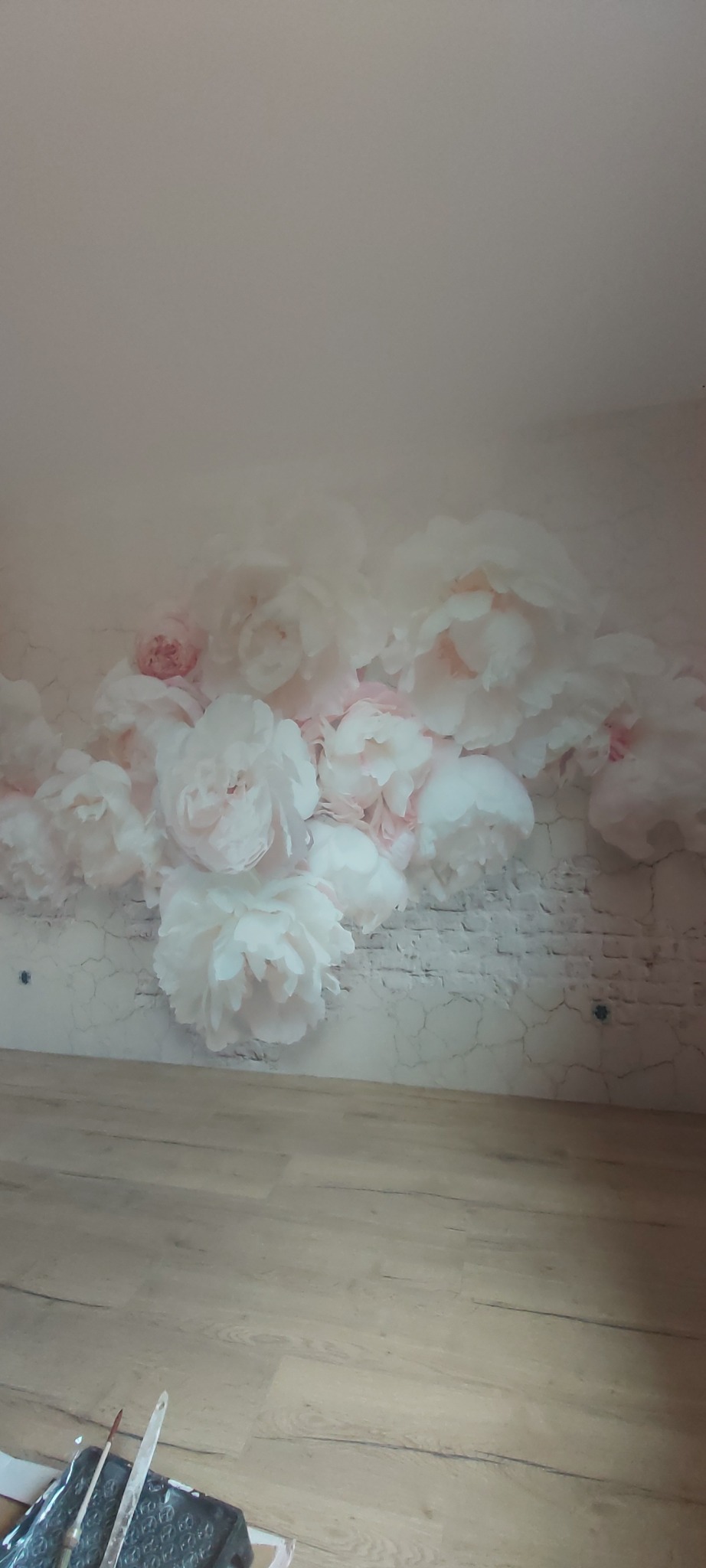 Fototapeta z motywem białych piwonii na ścianie imitującej cegłę, widoczna podłoga i narzędzia malarskie w rogu.