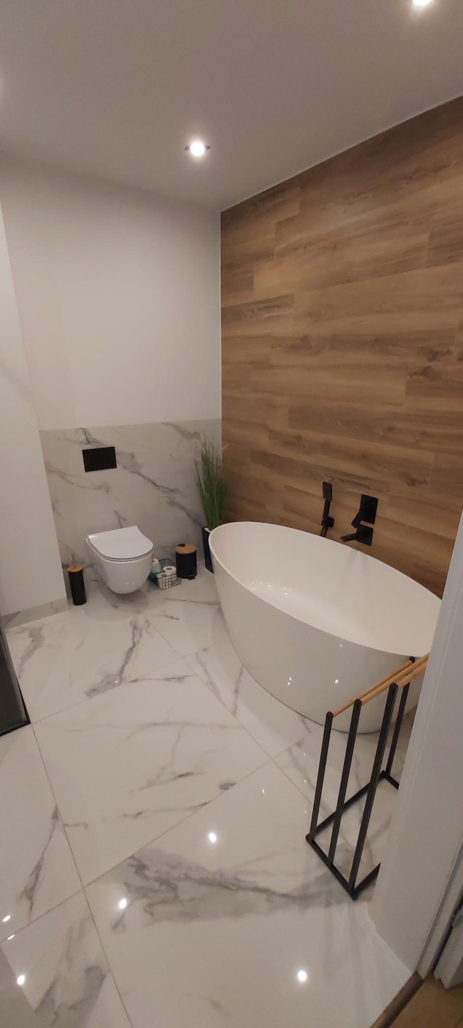 Nowoczesna łazienka z wolnostojącą wanną, marmurowymi płytkami i drewnopodobną okładziną ściany, minimalistyczna armatura w kolorze czarnym.