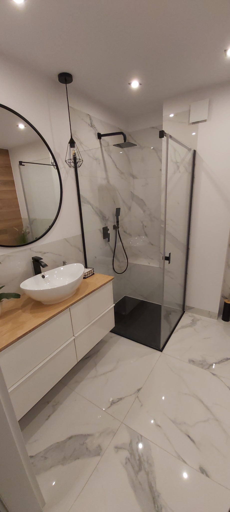 Nowoczesna łazienka z marmurowymi płytkami, kabiną prysznicową typu walk-in z czarnymi profilami, umywalką nablatową na szafce z drewnianym blatem i okrągłym lustrem z czarną ramą.