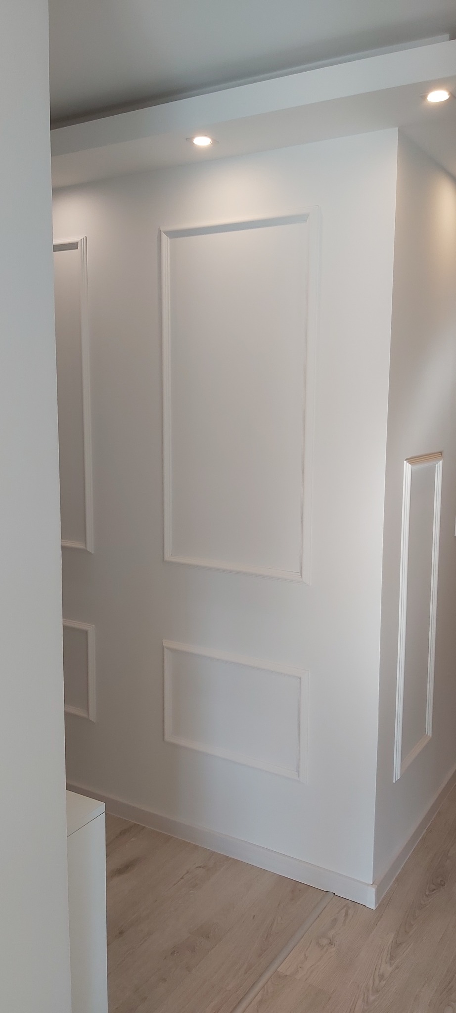 Jasne wnętrze z dekoracyjnymi, prostokątnymi listwami ściennymi, oświetleniem punktowym i podłogą z paneli w odcieniach beżu.