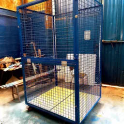 Niebieska, metalowa klatka magazynowa z kratką, na kółkach, z żółtą podłogą, widoczna w warsztacie, obok stół z narzędziami.