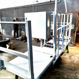 Wnętrze warsztatu z widoczną platformą roboczą ze stalową konstrukcją, białą blachą podłogową i skrzynką elektryczną, częściowo owiniętą folią, oraz mężczyzną robiącym zdjęcie w tle.