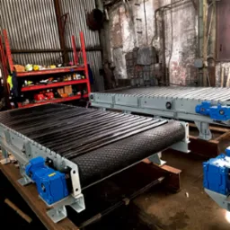 Dwa przemysłowe przenośniki taśmowe z niebieskimi silnikami w hali produkcyjnej, widoczne metalowe konstrukcje i elementy napędowe.