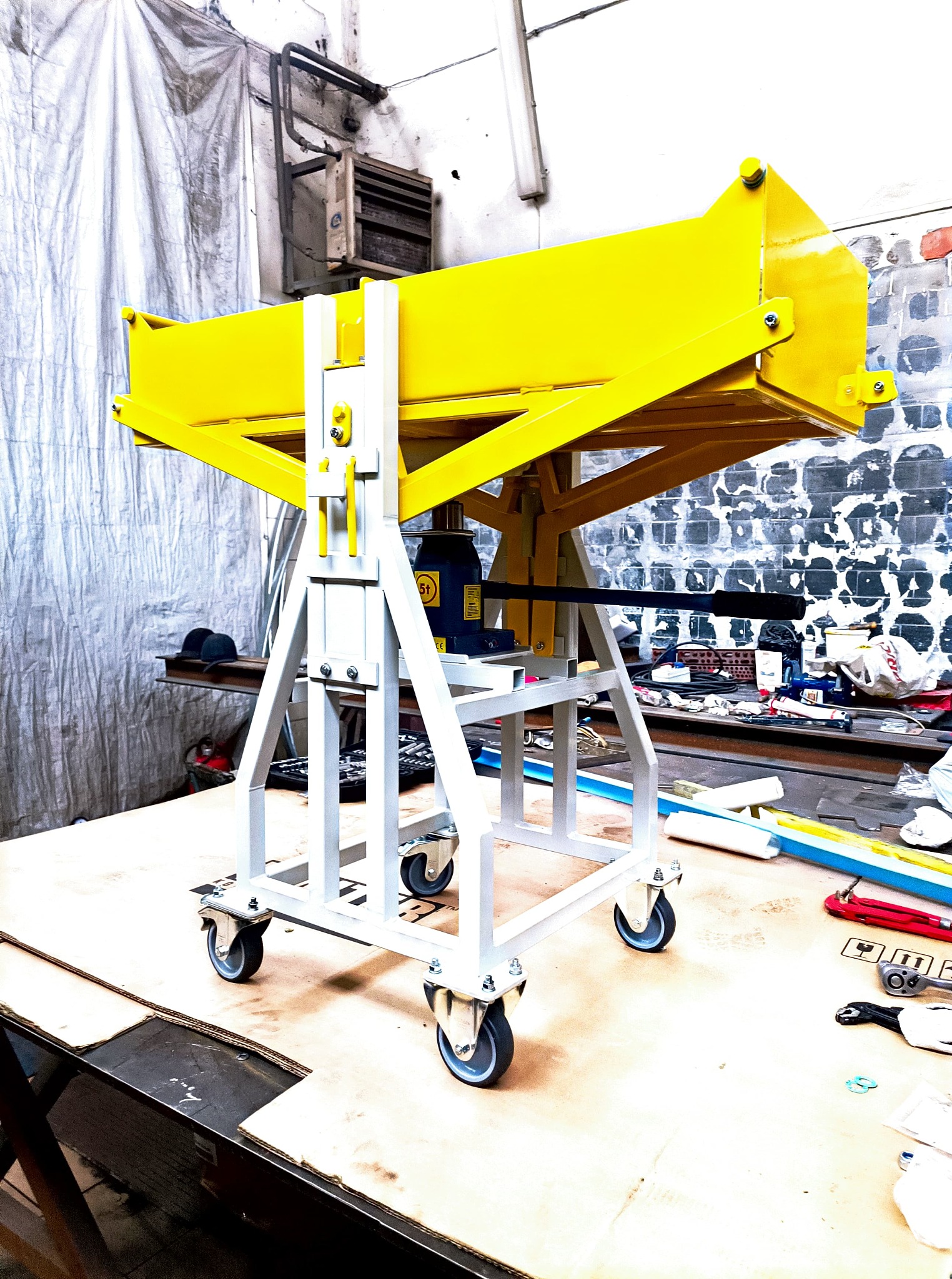 Żółto-biały, mobilny stojak z hydraulicznym podnośnikiem, prawdopodobnie do obróbki metalu, na tle warsztatu z narzędziami.